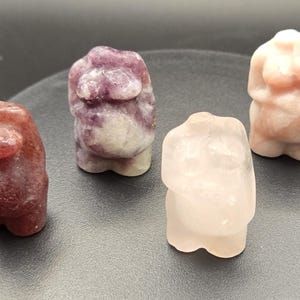 Può includere: Quattro figurine in pietra scolpita nei toni del rosso, viola e rosa. Le figure sono stilizzate, con forme arrotondate che suggeriscono un busto. Le pietre hanno una finitura lucida e le figure sono esposte su una superficie grigio scuro.