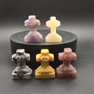 Può includere: Sei piccole sculture in pietra a forma di croce in vari colori, tra cui viola, giallo, grigio e rosso-marrone. Le sculture sono esposte su una superficie nera su sfondo scuro. Le croci hanno un aspetto dettagliato e strutturato.