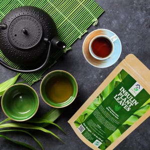 May include: Overhead shot of a tea set with a black cast iron teapot, two green tea cups, and a teacup with a blue rim. A package of Insulin Plant Leaves is also present. The tea is set on a dark gray surface with a green bamboo mat.