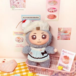 May include: A plush doll with a cartoon face, wearing a light blue dress and a white beret with a cherry on top, sits in a pink wire basket. The doll is surrounded by decorative cards and small toy food items. The text "SPRING SPRING" is visible.