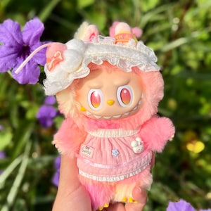 May include: A plush toy with a pink and yellow color scheme, featuring a doll-like face with large red eyes and a grumpy expression. The toy is wearing a pink dress with a white lace trim and a white bonnet. The text "Sweet" is visible.