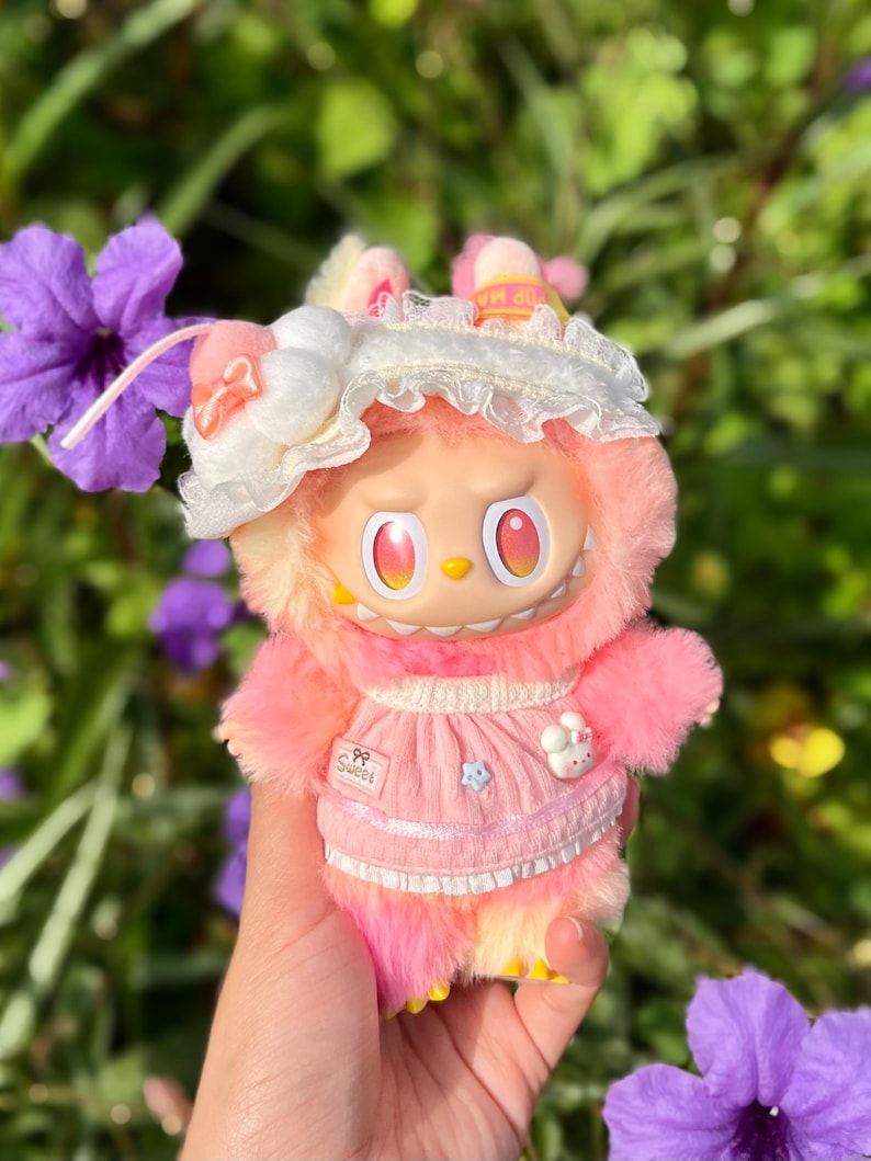 May include: A plush toy with a pink and yellow color scheme, featuring a doll-like face with large red eyes and a grumpy expression. The toy is wearing a pink dress with a white lace trim and a white bonnet. The text "Sweet" is visible.