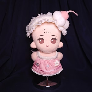 May include: A plush doll with a light pink face, large brown eyes, and a sweet expression. It wears a pink dress with a white lace trim and a matching headband with a cherry on top. The doll is displayed on a metal stand.
