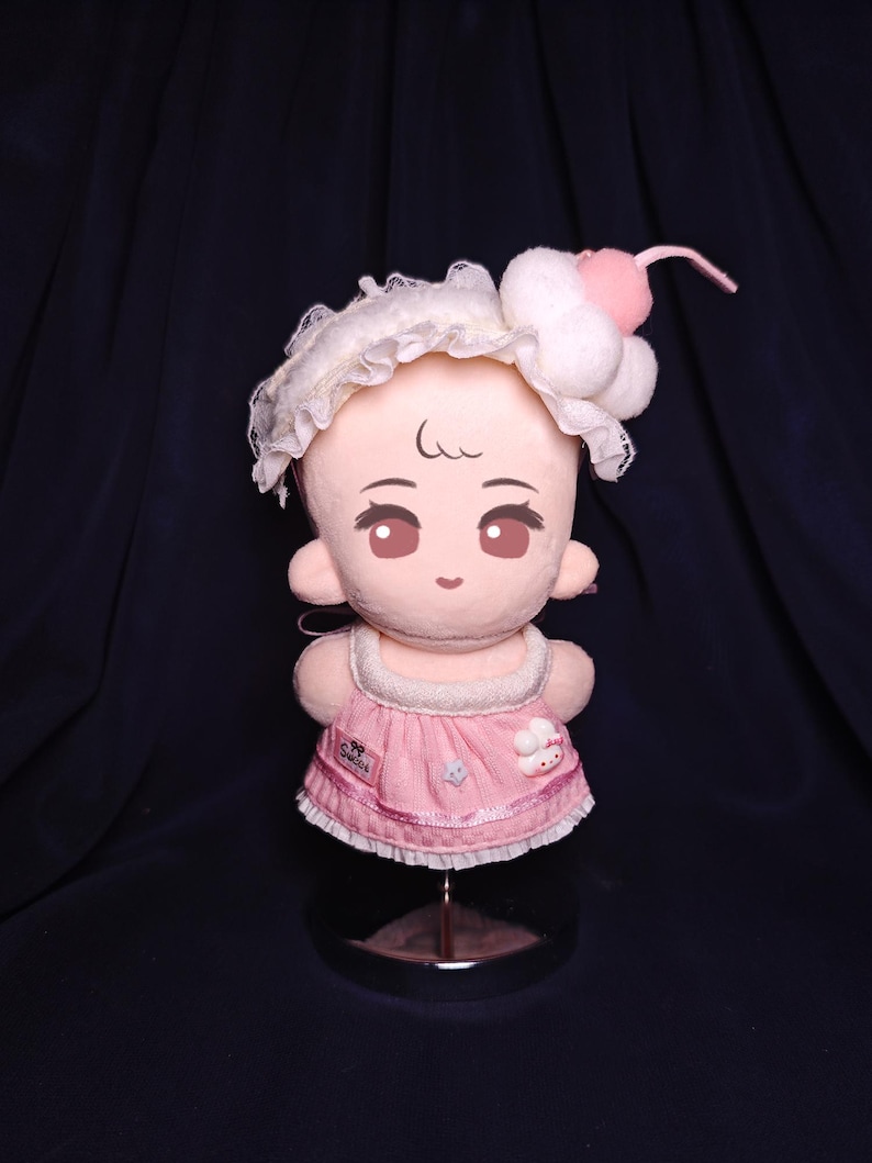 May include: A plush doll with a light pink face, large brown eyes, and a sweet expression. It wears a pink dress with a white lace trim and a matching headband with a cherry on top. The doll is displayed on a metal stand.