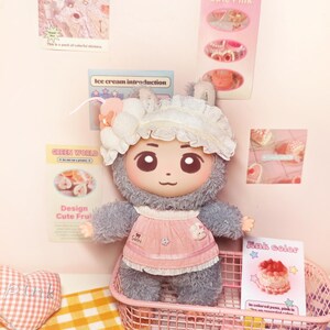 May include: A plush doll with a cute face, wearing a pink apron and a white bonnet. The doll is in a pink wire basket on a yellow and white checkered surface. The background includes posters with text like "Design Cute Pink" and "Ice cream introduction."