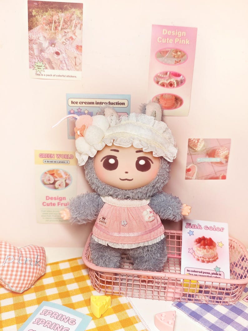 May include: A plush doll with a cute face, wearing a pink apron and a white bonnet. The doll is in a pink wire basket on a yellow and white checkered surface. The background includes posters with text like "Design Cute Pink" and "Ice cream introduction."