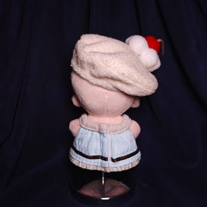 May include: A plush doll wearing a cream-colored hat with white pom-poms and a red accent. The doll has a light blue dress with brown trim. The doll is displayed on a clear stand against a dark blue background.