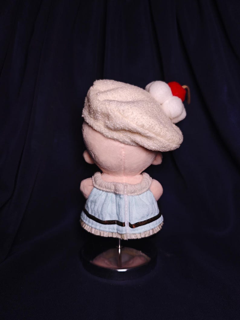 May include: A plush doll wearing a cream-colored hat with white pom-poms and a red accent. The doll has a light blue dress with brown trim. The doll is displayed on a clear stand against a dark blue background.