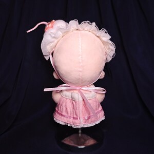 May include: A handmade pink doll with a bonnet and dress. The doll's head is a soft pink color, with a white lace-trimmed bonnet and a pink ribbon. The dress is a matching pink, with a ribbon tied at the back. The doll is displayed on a clear stand.