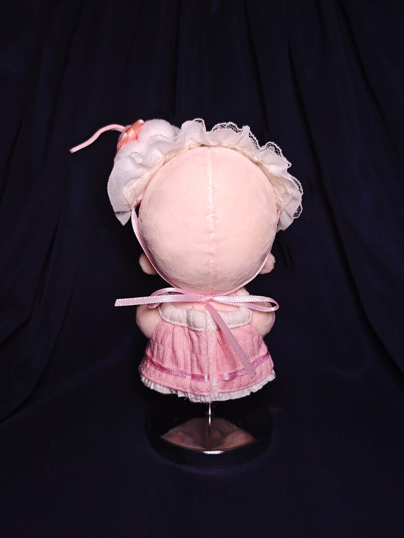 May include: A handmade pink doll with a bonnet and dress. The doll's head is a soft pink color, with a white lace-trimmed bonnet and a pink ribbon. The dress is a matching pink, with a ribbon tied at the back. The doll is displayed on a clear stand.