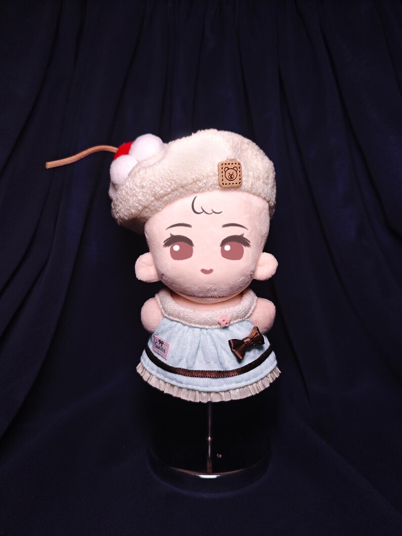 May include: A plush doll with a cream-colored hat adorned with a red and white cherry. The doll has a light blue dress with a brown bow and trim. The doll has a cute face with large eyes.