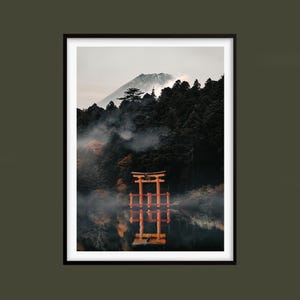 Puede incluir: Impresión enmarcada de un paisaje japonés. La imagen muestra una vibrante puerta torii naranja reflejada en agua tranquila, con un bosque brumoso y una montaña al fondo. La obra de arte está situada sobre un fondo oscuro y neutro.