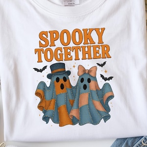 May include: White t-shirt with the text "SPOOKY TOGETHER" in orange. The shirt features two ghost characters, one with a hat and the other with a bow, in blue and orange patchwork. Small black bats are also included in the design.