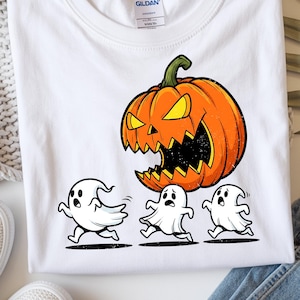 Puede incluir: Camiseta blanca con un diseño de Halloween. El gráfico muestra una gran calabaza naranja con una expresión amenazante persiguiendo a tres fantasmas de dibujos animados. El diseño es de estilo caricatura.