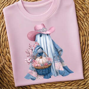 Puede incluir: Sudadera rosa claro con una ilustración caprichosa de un fantasma con un sombrero de vaquero rosa con una banda plateada. El fantasma lleva una chaqueta vaquera y sostiene una cesta con una calabaza y flores. El diseño está sobre un fondo rosa claro.
