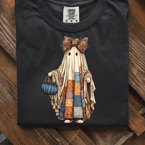 Könnte beinhalten: Schwarzes T-Shirt mit einem skurrilen Geisterdesign. Der Geist trägt eine Blumen-Schleife und einen Patchwork-Schal und hält eine blaue, kürbisförmige Tasche. Das Shirt hat einen Rundhalsausschnitt und das Etikett lautet "Comfort Colors".