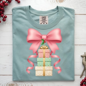 Op de afbeelding: Lichtblauw T-shirt met een ontwerp van drie gestapelde cadeau dozen, een grote roze strik en linten. De dozen zijn roze, mintgroen en crème, met gouden details. Een takje hulst bevindt zich in het midden van de strik.