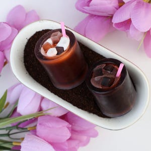 May include: Two iced coffee candles with pink straws sit in a white oval dish filled with coffee grounds. The candles have ice cubes and cream. Pink tulips surround the dish on a white surface.