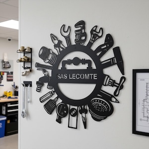 Peut inclure: Décoration murale en métal noir représentant un cercle orné d'outils de plomberie. Le texte "SAS LECOMTE" est inscrit au centre. Des accessoires de plomberie supplémentaires sont fixés au mur.