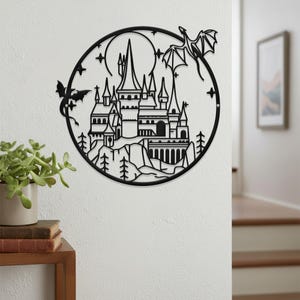 May include: Black metal wall art depicting a castle scene within a circular frame. The design includes a castle with multiple turrets, trees, mountains, a full moon, stars, and two dragons. The art is mounted on a white wall.