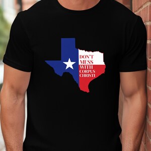 May include: Black t-shirt featuring a Texas state outline in blue, white, and red, with a white star. The text "DON'T MESS WITH CORPUS CHRISTI" is printed on the right side of the Texas outline.