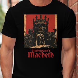 May include: A black t-shirt featuring a graphic print of a large skull wearing a crown above a figure holding a sword. The design is set against a red background with dark, distressed details. Below the illustration, the text reads Shakespeare's Macbeth.