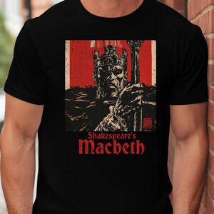 Peut inclure: Un t-shirt noir à manches courtes avec une impression graphique représentant une figure couronnée tenant un sceptre sur fond rouge. Le texte "Shakespeare's Macbeth" est imprimé en rouge sous l'illustration dans une police serif stylisée.
