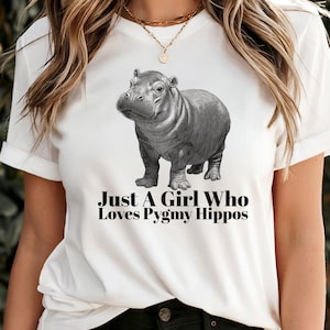 May include: White t-shirt with a grayscale illustration of a pygmy hippo. The text "Just A Girl Who Loves Pygmy Hippos" is printed below the hippo. The shirt has short sleeves and a crew neck.