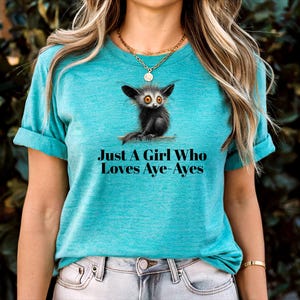 May include: Teal t-shirt with an aye-aye illustration, a nocturnal lemur, on a branch. The text "Just A Girl Who Loves Aye-Ayes" is printed below. The shirt has rolled-up sleeves. The tee is a vibrant teal colour.