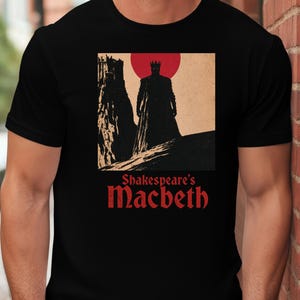 Peut inclure: Un t-shirt noir à manches courtes avec une impression graphique représentant un roi en silhouette devant un grand soleil rouge et un château. Sous l'illustration, le texte "Shakespeare's Macbeth" est écrit en police gothique rouge.