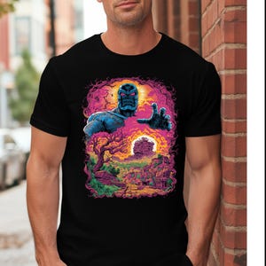 Op de afbeelding: Zwart t-shirt met een kleurrijke afbeelding. Het ontwerp toont een blauwhuidig ​​personage met een uitgestrekte hand, tegen een levendig landschap met een zonsondergang en ruïnes, omlijst door een roze en paarse rand.