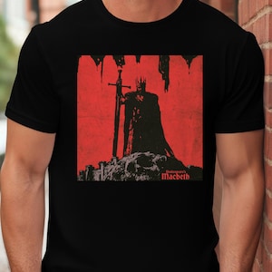Peut inclure: Un t-shirt noir à manches courtes avec une impression graphique représentant une silhouette couronnée tenant une épée sur fond rouge. Le motif inclut le texte Shakespeare's Macbeth en lettres rouges en bas à droite de l'illustration.