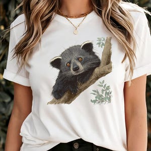 Op de afbeelding: Wit T-shirt met een gedetailleerde illustratie van een zwarte en grijze binturong op een tak. De binturong heeft bruine ogen en is omgeven door groene bladeren. Het shirt heeft korte mouwen.