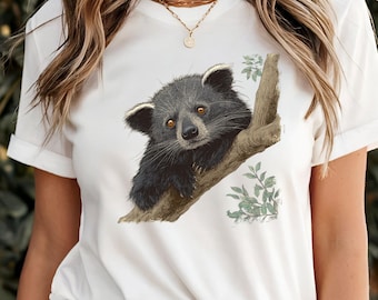 May include: White t-shirt with a detailed illustration of a black and grey binturong on a tree branch. The binturong has brown eyes and is surrounded by green leaves. The shirt has short sleeves.