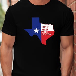 Może przedstawiać: Czarny t-shirt z konturem stanu Teksas w kolorze niebieskim, białym i czerwonym, białą gwiazdą i napisem "DON'T MESS WITH DENTON". Koszulka ma okrągły dekolt.