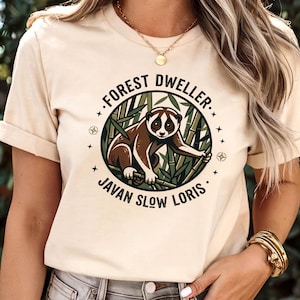 Peut inclure: Un t-shirt couleur crème avec un motif circulaire représentant un loris lent de Java brun et blanc assis parmi des tiges de bambou vertes. Le texte "FOREST DWELLER" est arqué au-dessus de l'animal et "JAVAN SLOW LORIS" est inscrit sous le cercle.