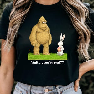 May include: Black t-shirt featuring a cartoon illustration of a large, tan gorilla and a white rabbit holding an Easter egg. The text "Wait...you're real??" is printed below the characters.