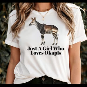 May include: White t-shirt with an okapi illustration, a mammal with a zebra-like pattern on its legs. The text "Just A Girl Who Loves Okapis" is printed below the okapi image. The t-shirt is white.