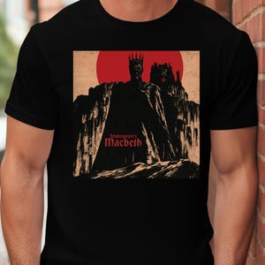 Może przedstawiać: Czarny t-shirt z krótkim rękawem z nadrukiem graficznym przedstawiającym ukoronowaną postać w pelerynie stojącą przed zamkiem i dużym czerwonym słońcem. Napis "Shakespeare's Macbeth" jest wydrukowany czerwoną czcionką w lewym dolnym rogu grafiki.