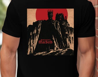 May include: A black short-sleeved t-shirt featuring a graphic print of a crowned figure in a cape standing before a castle and a large red sun. The text "Shakespeare's Macbeth" is printed in red lettering on the lower left side of the graphic.