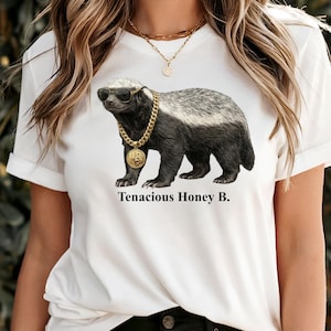 Op de afbeelding: Een wit t-shirt met een afbeelding van een honingdas die een zwarte zonnebril en een dikke gouden ketting met een ronde hanger draagt. Onder het dier staat de tekst Tenacious Honey B. in een zwart schreeflettertype. T-shirt met korte mouwen.