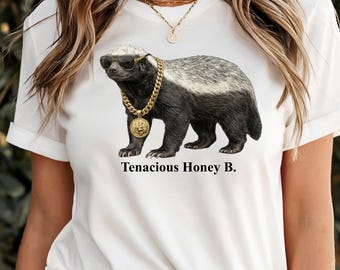 May include: A white t-shirt featuring a graphic of a honey badger wearing black sunglasses and a thick gold chain with a circular pendant. Below the animal, the text reads Tenacious Honey B. in a black serif font. The shirt has short sleeves.