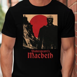 Peut inclure: Un t-shirt noir à manches courtes avec une impression graphique représentant une silhouette couronnée devant un grand soleil rouge et une falaise sombre. Sous l'illustration, le texte Shakespeare's Macbeth est écrit en rouge.