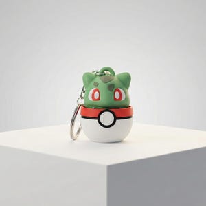 Puede incluir: Un llavero con figura de Bulbasaur verde sobre una Poké Ball blanca y roja. El Bulbasaur tiene ojos rojos y una cabeza verde. La Poké Ball es blanca con una franja roja y un círculo blanco y negro en el centro.