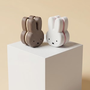 May include: Two rabbit-shaped objects, one brown and one white, each featuring a central vertical seam, two upright ears, and a face with two black dot eyes and a black cross-shaped mouth. The figures have a smooth, layered texture.
