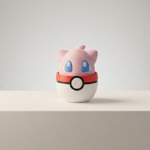 以下が含まれることがあります： 青い目のピンクと白の漫画のキャラクターのフィギュアが、赤と白のボールの上にあります。フィギュアは丸みを帯びた体と大きな、表情豊かな目をしています。ボールの中央には黒い円があります。