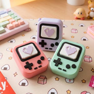 以下が含まれることがあります： パステルカラーの携帯ゲーム機型アクセサリー3点。ピンク、紫、緑のデバイスは、画面にハートのデザインと黒い十字キーが付いています。背景にはキーボード、お菓子、小さなフィギュアなどがあります。