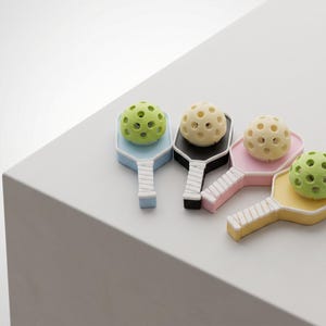 Puede incluir: Cuatro mini sets de pickleball en colores pastel: azul claro, negro, rosa y amarillo. Cada set incluye una pala y una pelota perforada verde o crema. Las palas tienen mangos blancos.