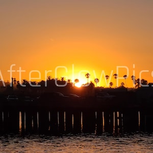 May include: A sunset scene with a silhouetted pier and water in the foreground. The sky is a gradient of orange and yellow, with the sun's glow visible. Palm trees and buildings are silhouetted against the bright sky. The text "AfterGlowPics" is visible.