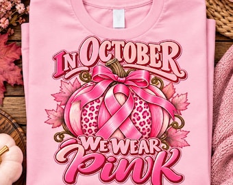 May include: A light pink t-shirt featuring a graphic of a pumpkin with a pink ribbon and leopard print accents. The text reads "IN OCTOBER WE WEAR PINK" in bold pink lettering. The design includes pink maple leaves and a pink bow on the pumpkin.